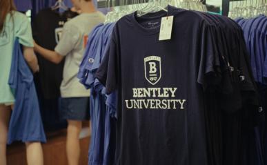 A rack of navy blue t-shirts with the Bentley logo, with visible price tag. In the background are two students, their backs to camera, browsing other items.