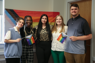Five members of the inaugural class of Rainbow Scholars pose in front of a rainbow flag. Two also hold smaller rainbow flags in their hands.