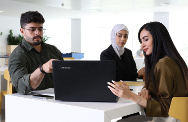 Three Kuwait College of Science and Technology students, one male and two female, sit together at a white table and look toward an open laptop computer.