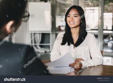 Woman with long black hair holding paper and sitting at a table with an interviewer across from her
