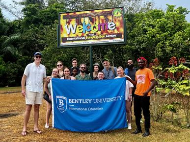 Bentley students and their group leaders pose with a blue Bentley banner and their local tour guide at the Stingless Bee Centre in Ghana.