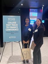 Two female students in business attire stand next to a conference sign that reads "College Fast Track to Finance Conference"