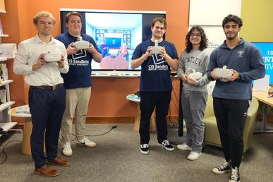 Five Bentley students and CIS Sandbox tutors — Colin Minarik, Alden Shoemaker, Vincent Paratore, Andre Noonan and Lucas Fernandez de Assis — pose with VR Headsets in front of a giant monitor displaying a Bodyswaps module.