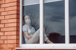 Young woman with face mask looks out her window during COVID-19 quarantine.
